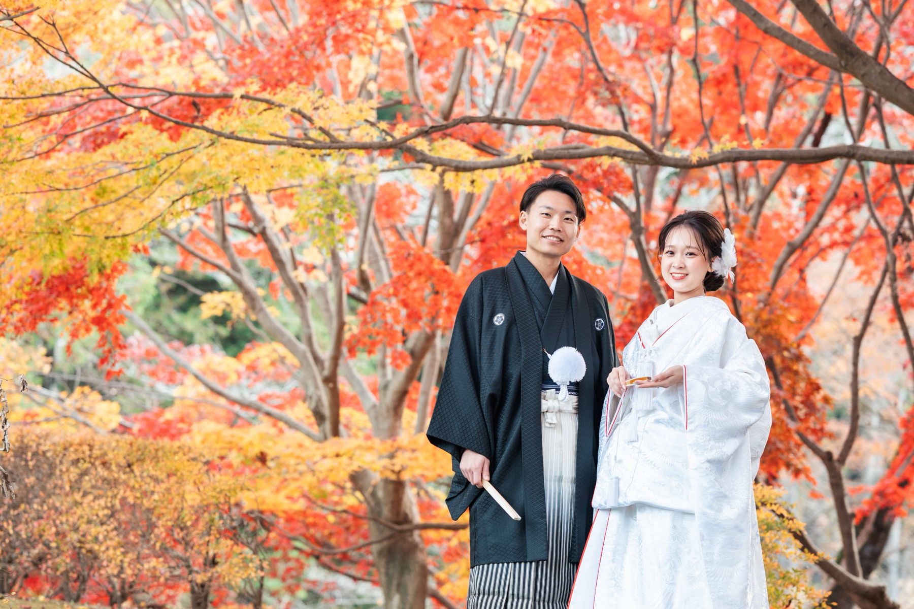 紅葉の下に立つ紋付袴と白無垢のふたり