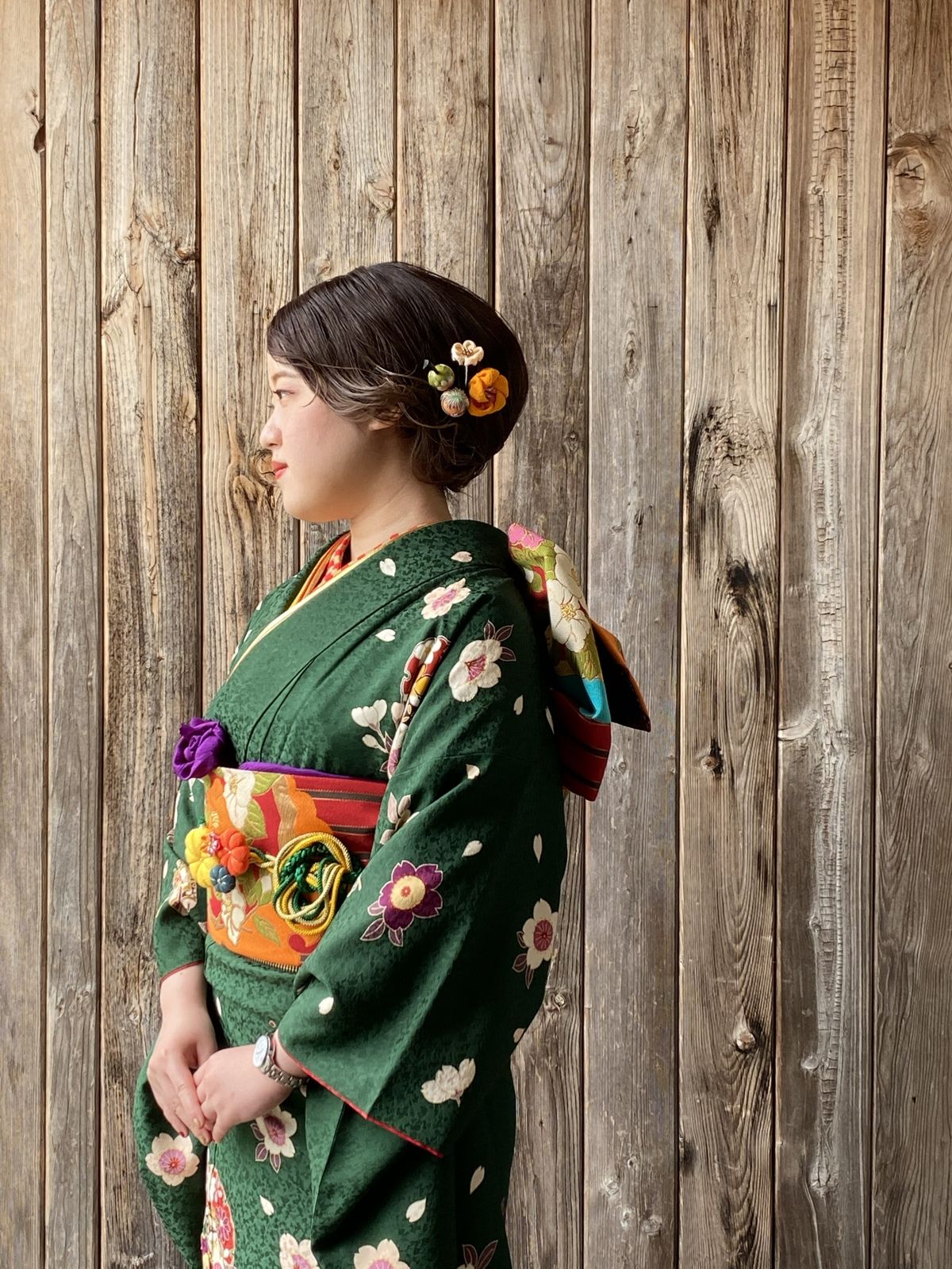 杉板の壁の前に立つ緑の振袖姿の女性の横顔
